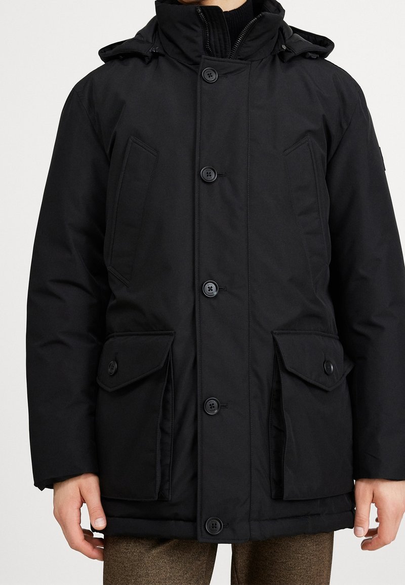 Černá izolovaná parka s vysokým límcem, zapínáním na knoflíky a dvěma velkými předními kapsami. Má odnímatelnou kapuci a hladkou texturu.