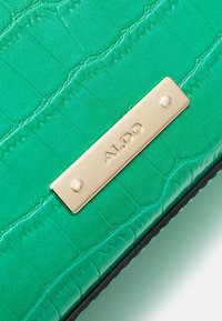 Surface en cuir vert texturé avec une plaque métallique rectangulaire dorée embossée du nom de la marque ALDO.
