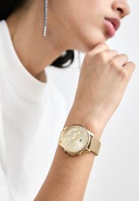 Montre-bracelet en or avec un bracelet en mailles, cadran rond, face crème claire et sous-cadrans. Positionnée sur un poignet portant une chemise blanche.