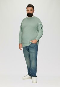 Lichtgroene sweatshirt met structuur en een witte kraag, met een patch pocket op de mouw. Draagt in combinatie met blauwe jeans en witte sneakers.