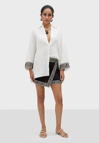 Camicia bianca di lino con chiusura a bottoni, gonna nera con bordi a stampa zebra. Il modello indossa accessori dorati e sandali bassi.