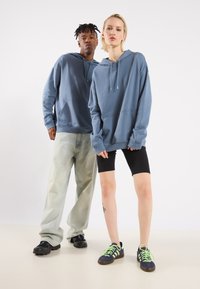 Due modelli indossano felpe blu con coulisse. Un modello ha dei jeans chiari, mentre l'altro indossa pantaloni corti neri e scarpe con accenti verde neon.