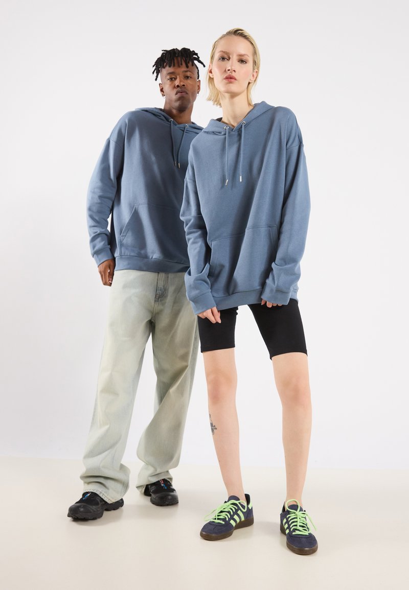 Due modelli indossano felpe blu con coulisse. Un modello ha dei jeans chiari, mentre l'altro indossa pantaloni corti neri e scarpe con accenti verde neon.