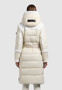 Nadýchaný krémově zbarvený kabát s bílým kožichem na límci, páskem v pase a logem na zádech. Texturovaná látka s prošívanými částmi.