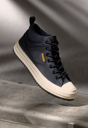 Černý kotníkový tenisky s bílou gumovou špičkou a podrážkou, černými šněrovadly, žlutou štítkem „Voděodolný“, vznášející se nad betonovým povrchem.