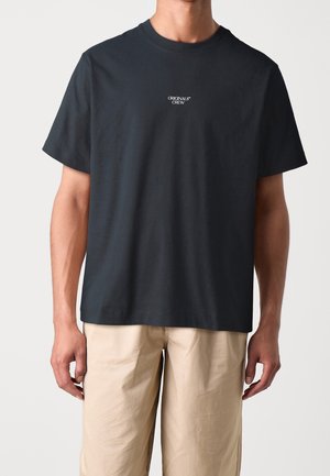 T-shirt noir à manches courtes avec col rond, portant un petit texte blanc "ORIGINALS CREW" sur la poitrine, associé à un pantalon beige sur une personne debout.