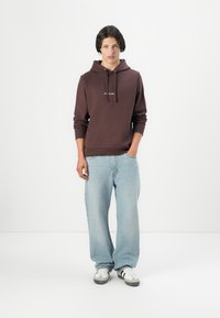 Dark brown hoodie with a front pocket, paired with light blue wide-leg jeans and black and white sneakers. Plain background.