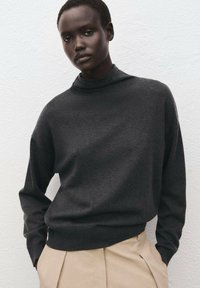 Temno sivi pulover z visokim ovratnikom in dolgimi rokavi, narejen iz mehkega, teksturiranega materiala. Kombiniran z bež hlačami po meri.