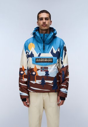 Uomo che indossa una giacca blu, bianca e marrone con stampe di montagne e foreste, logo Napapijri e pantaloni beige, in piedi su uno sfondo semplice.