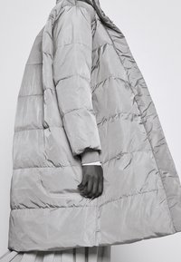 Polstret grå frakke med en lang silhuet, der har horisontale sømme, en høj krave og ribbede manchetter, vist på en person i profil.