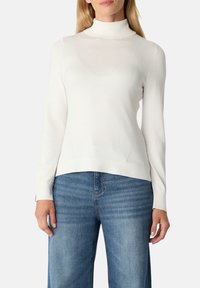 Femme portant un pull à col roulé à manches longues blanc et un jean en denim bleu devant un fond uni.
