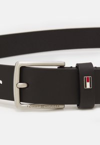 Svart läderbälte med silvermetallspänne. Har präglad "TOMMY HILFIGER" och en liten flerfärgad logotypaccent nära spännet.