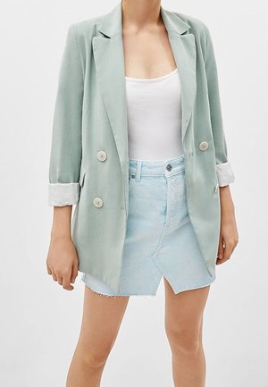 Blazer de doble botonadura verde claro con mangas enrolladas, combinado con una camiseta sin mangas blanca y una falda de mezclilla azul claro con dobladillo deshilachado.