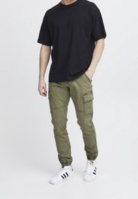 Zwart T-shirt met korte mouwen, olijfgroene cargobroek met meerdere zakken, elastische enkelboorden en witte sneakers met zwarte strepen.