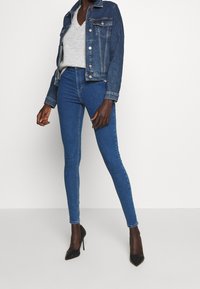 Tummansininen farkkutakki hopeisilla napilla ja taskuilla, yhdistettynä korkeavyötäröisiin sinisiin pillifarkkuihin. Mustat korkokengät täydentävät asun.