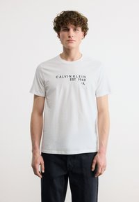 Νέος άνδρας με σγουρά μαλλιά, ντυμένος με λευκό T-shirt Calvin Klein με μαύρο κείμενο, σε συνδυασμό με σκούρο παντελόνι, μπροστά σε μονόχρωμο φόντο.