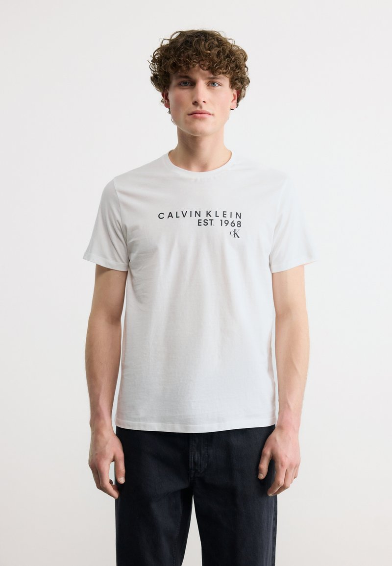 Νέος άνδρας με σγουρά μαλλιά, ντυμένος με λευκό T-shirt Calvin Klein με μαύρο κείμενο, σε συνδυασμό με σκούρο παντελόνι, μπροστά σε μονόχρωμο φόντο.