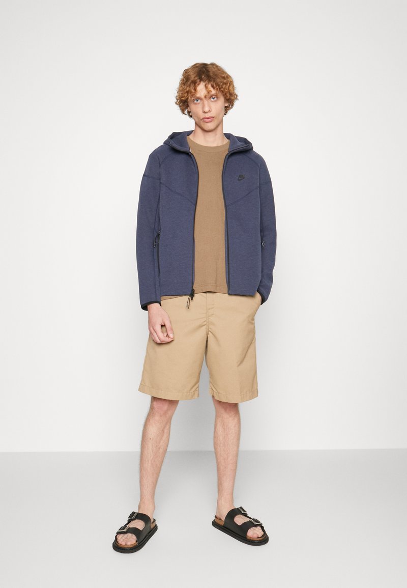 Sweat à zip bleu marine avec capuche, t-shirt beige, shorts beiges et sandales noires avec boucles. Texture de tissu lisse, coupe décontractée et design simple.