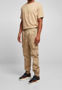 Beige t-shirt i bomull med korta ärmar och avslappnad passform, kombinerad med beiga cargo-byxor med sidofickor och elastisk midja. Mörka sneakers.