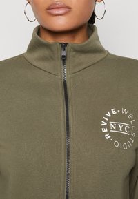 Veste zippée vert olive avec col haut, fermeture éclaire noire et logo circulaire blanc portant le texte "REVIVE - WELL STUDIO NYC" sur le côté gauche.