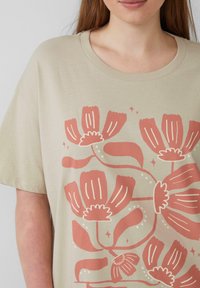 Beżowa koszulka z kwiatowym wzorem w kolorze koralowym i białym. Posiada krótkie rękawy oraz okrągły dekolt. Gładka faktura.