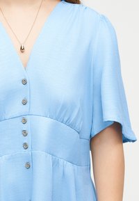 Γυναίκα που φοράει ανοιχτό μπλε φόρεμα με κουμπιά και κοντά μανίκια, με χρυσό κολιέ με σταγόνα σε λεπτή αλυσίδα.