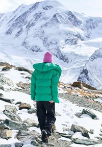 Osoba v zelenej bunde a ružovej čiapke kráča po skalnatom, zasneženom horskom teréne s veľkými zasneženými vrcholmi v pozadí.