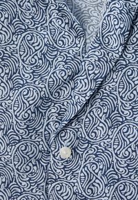 Tissu à motifs bleus et blancs présentant des tourbillons et des courbes, avec un petit bouton rond blanc. Texture lisse, design léger.