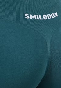 Teal sportleggings met een geribde tailleband waarop de merknaam "SMILODOX" in witte tekst staat. Close-up van de stoftextuur.