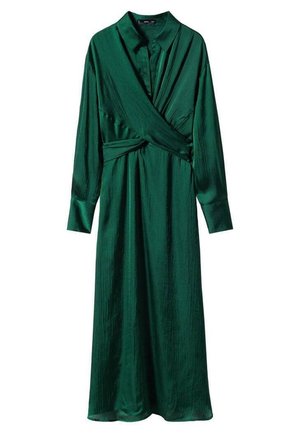 Femme portant une robe portefeuille vert foncé à manches longues avec un col et des sandales à talons noires à brides, debout sur un sol clair contre un mur blanc.