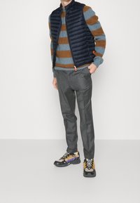 Marinestribet quiltet vest over en blå og brun stribet sweater, parret med grå bukser og multicolor sportssko.