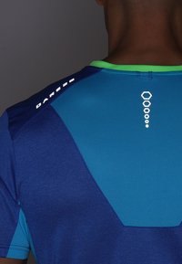 Camiseta deportiva azul con un cuello verde. Presenta un bloqueo de color dinámico con tejidos texturizados y un detalle de logotipo reflectante en la parte posterior.