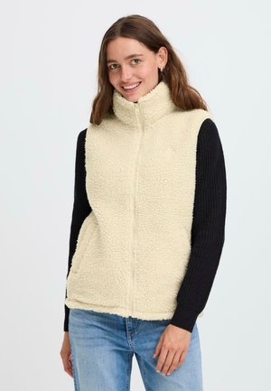 Vrouw in een crèmekleurige sherpa fleece vest over een zwarte gebreide trui met lange mouwen en blauwe spijkerbroek, glimlachend met ontspannen handen.