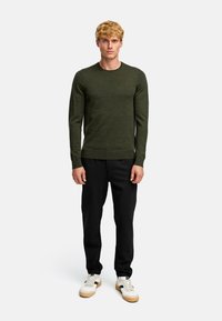 Junger Mann mit kurzen, lockigen blonden Haaren, der einen dunkelgrünen Langarmpullover, schwarze Hosen und weiße Sneakers mit beigefarbenen Sohlen trägt.