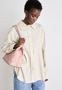Light beige oversized linen shirt with a pointed collar, button front. Model holds a soft pink quilted handbag with a curved shape and contrast trim.
