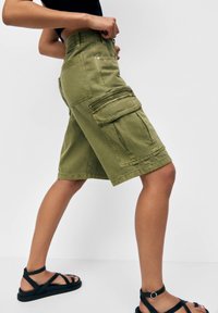 Person iført olivengrønne cargo shorts med store sidelommer og sorte sandaler med stropper, der står foran en ensfarvet baggrund.