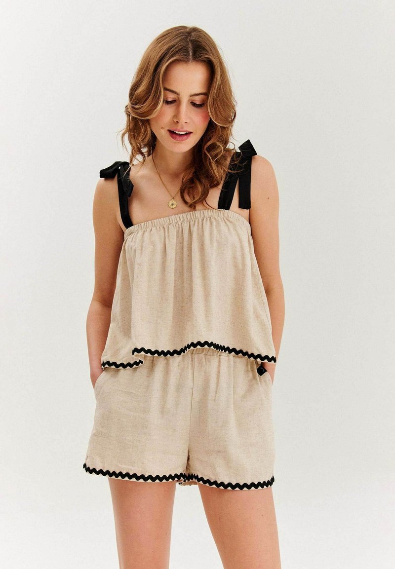 Béžový ľanový overál s volánovými vrstvami, čiernymi stužkami a vlnitými okrajmi. Obsahuje elastický pás a bočné vrecká.