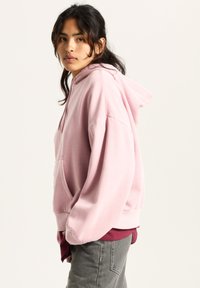 Pink hoodie with a front pocket and drawstring hood. Soft fabric, oversized fit, ribbed cuffs, layered over a maroon shirt, worn with gray pants.
