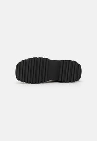 Semelle en caoutchouc noire avec de profondes rainures horizontales, surface texturée et bout arrondi. Design minimaliste avec un profil plat.
