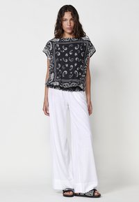 Blouse noire à motifs avec des manches courtes, ornée de motifs paisley blancs ; associée à un pantalon large blanc et des sandales noires à lanières.