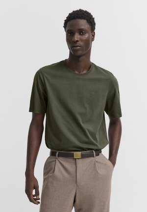 Mann trägt ein olivgrünes kurzärmliges T-Shirt, das in beige plissierte Hosen mit dunklem Gürtel gesteckt ist und vor einem schlichten Hintergrund steht.