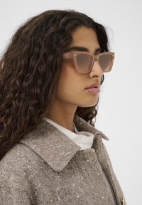 Očala svetlo rjave barve z rektangularno obliko in prozornim okvirjem. Oseba ima kodraste lase in nosi teksturirano rjavo oblačilo.