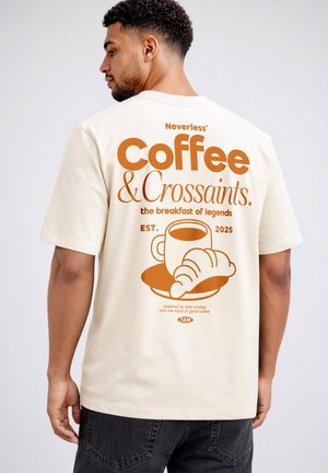 Man wearing beige T-shirt with orange graphic of coffee cup and croissant and text "Coffee & Crossaints, the breakfast of legends."