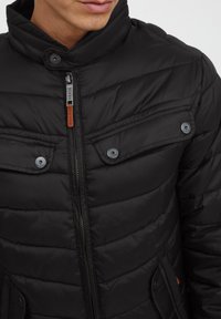 Black puffer jacket featuring a high collar, zip front, two chest pockets with button closures, and quilted texture.