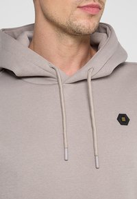 Lichtgrijze hoodie van zacht materiaal met een capuchon en trekkoord. Voorzien van een klein zwart hexagonaal logo met gele accenten.