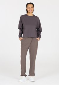 Dunkelgraues, kurz geschnittenes Langarmshirt, kombiniert mit lockeren braunen Hosen. Das Modell trägt weiße Sneaker. Einfaches Design, weiche Stofftextur.