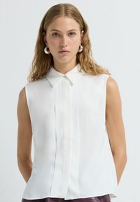 Weißes ärmelloses Bluse mit Kragen, Frontknopfverschluss und glattem, leichtem Stoff. Das Modell trägt große runde Ohrringe.