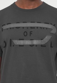 Γκρι βαμβακερό μπλουζάκι με έντονο γραφικό που γράφει "PROPERTY OF D.I.E." και δύο αντίθετες γκρι υφασμάτινες λωρίδες στο στήθος.