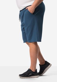 Pantaloni corti da uomo blu in felpa con vita elasticizzata e tasche laterali, abbinati a sneakers nere con suola grigia chiara e dettagli con il logo.