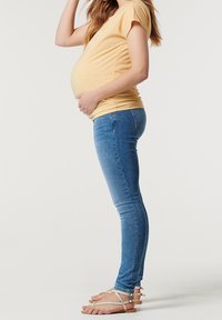 Těhotenský outfit skládající se z žlutého trička s krátkým rukávem a modrých skinny džínů, doplněný krémovými sandály na řemínky, zobrazený ze strany.